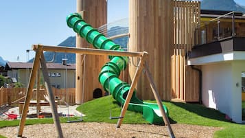 Ein Spielplatz mit einer grünen Rutsche und Schaukeln, umgeben von einer Rasenfläche und Holzelementen.
