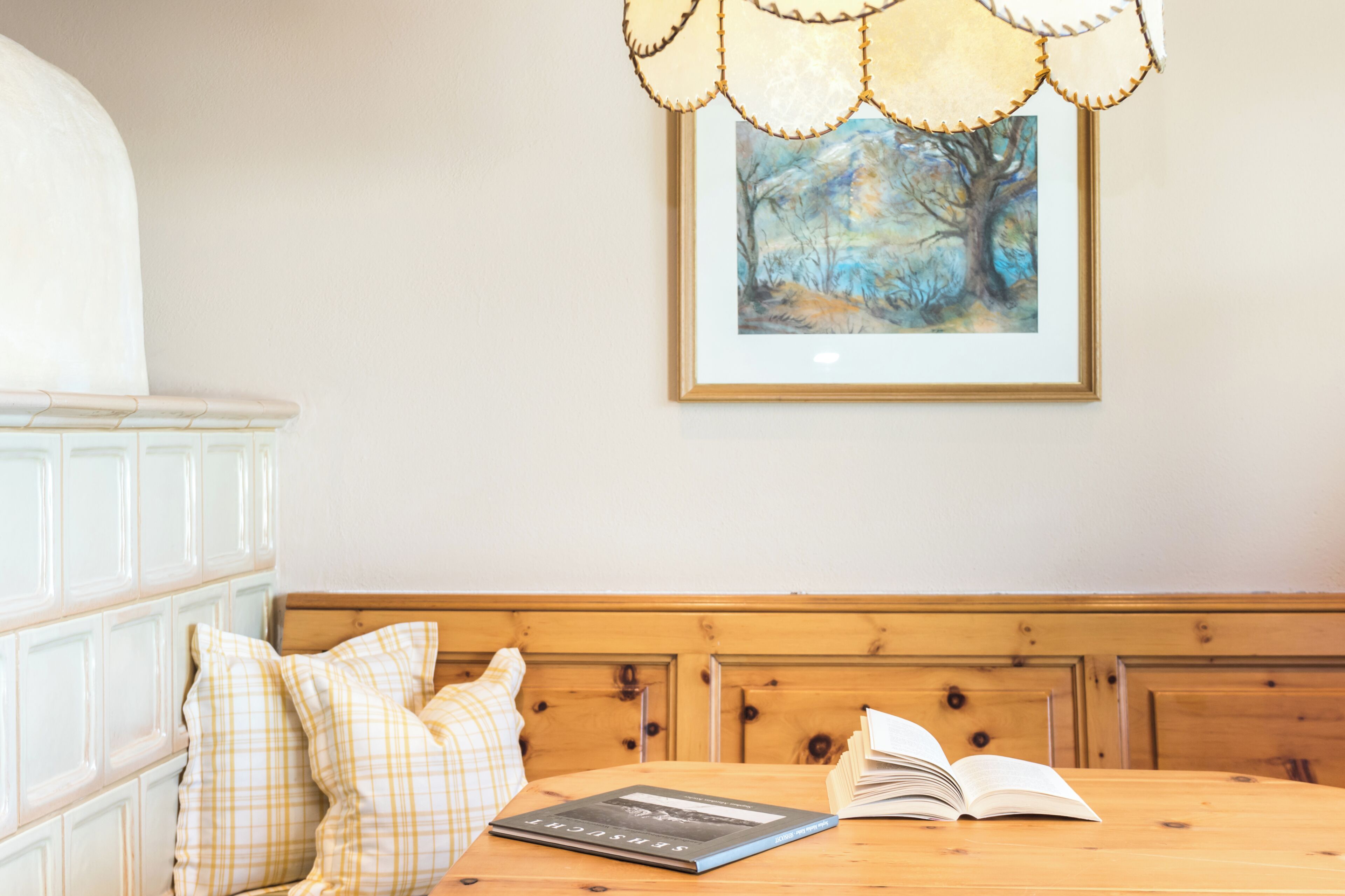 Una habitación con un banco de madera, una mesa con un libro abierto, un cuadro enmarcado en la pared y un armario blanco.