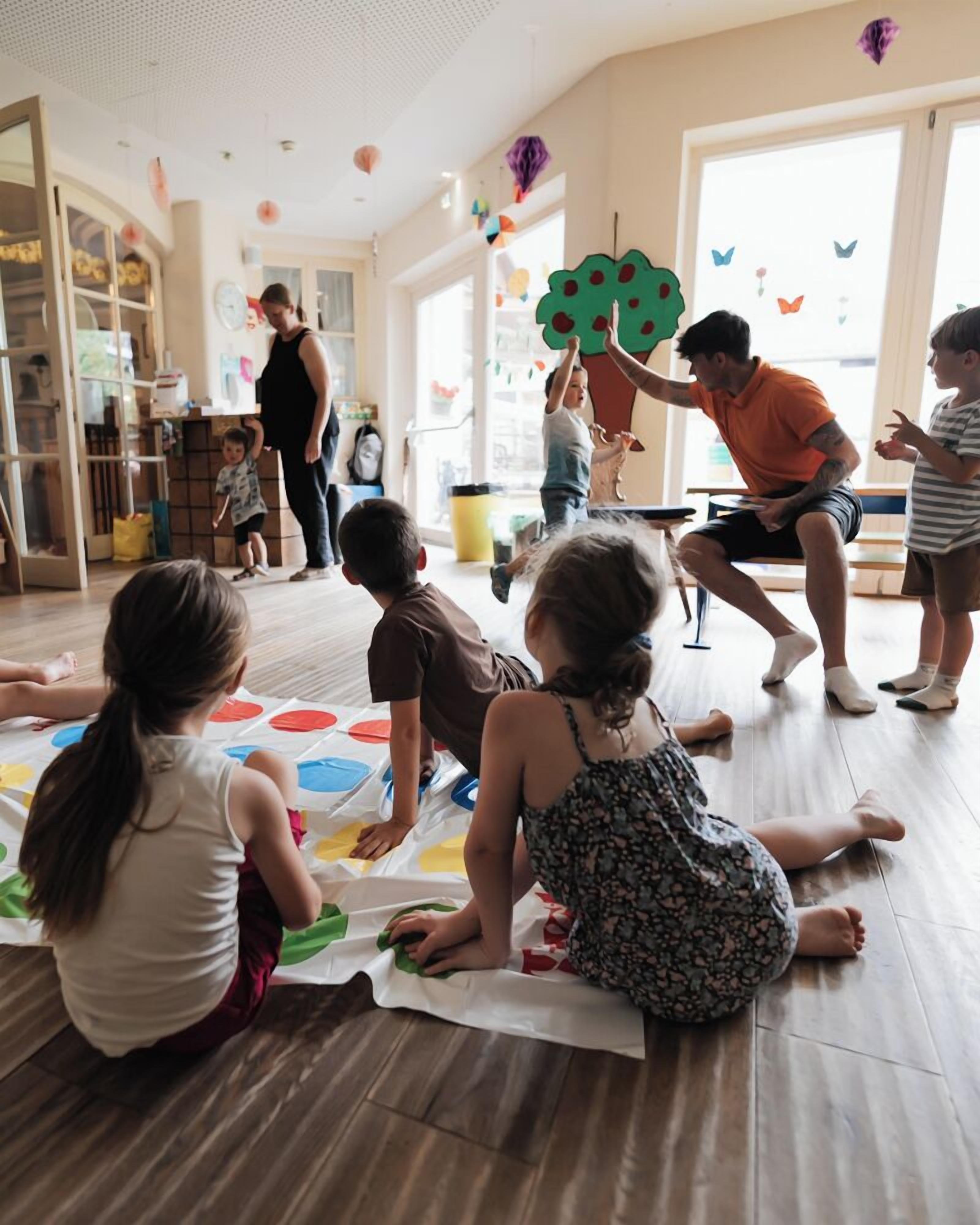A group of children engaged in a craft activity on a mat, with adults supervising in a room with large windows and a wooden floor.