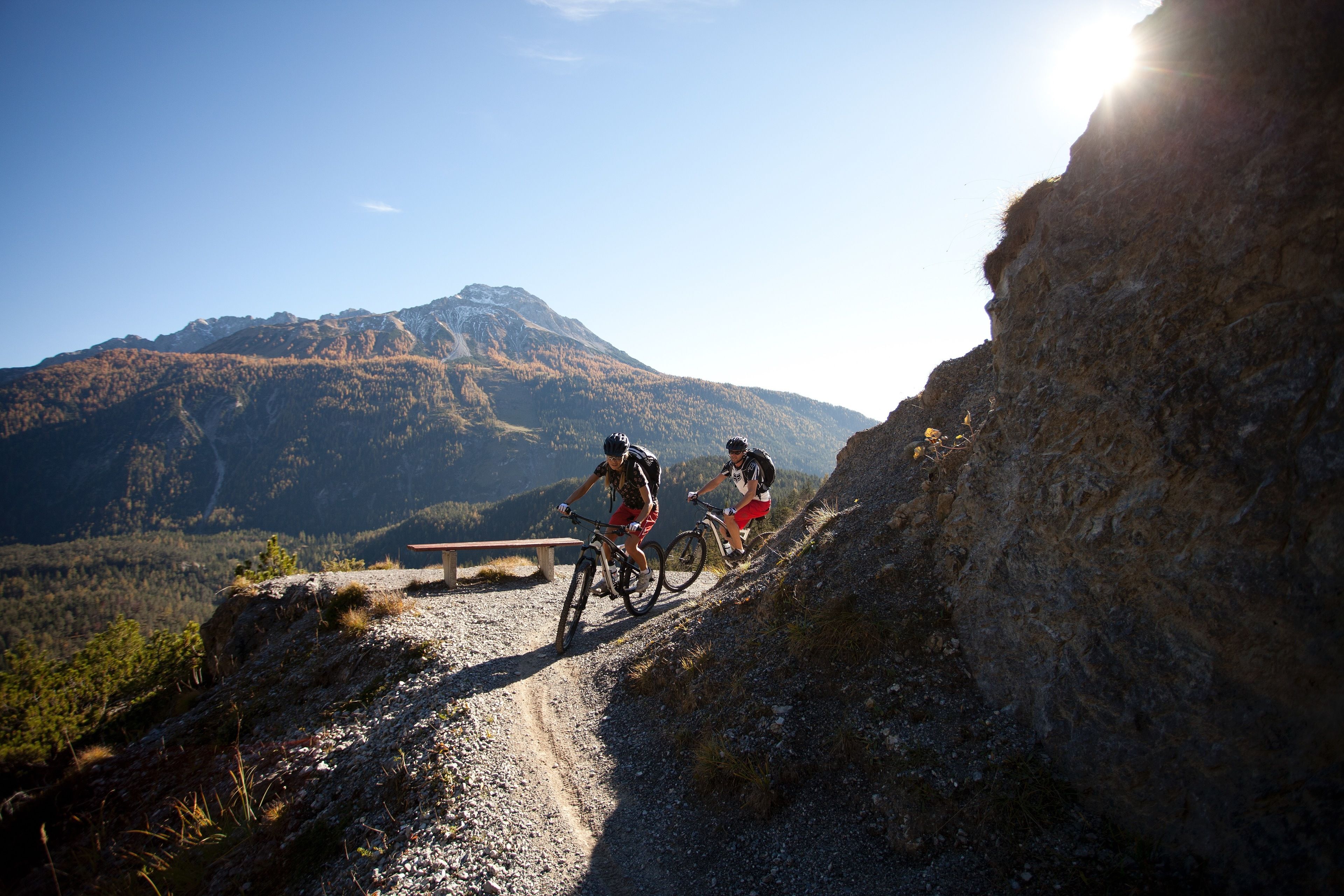 Two cyclists on a mountain trail with a bench and a picnic table.