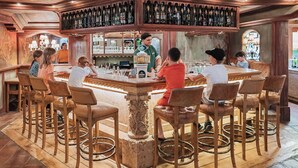 Uma área de bar com uma mesa redonda central rodeada por cadeiras, um barman e uma adega acima do balcão.