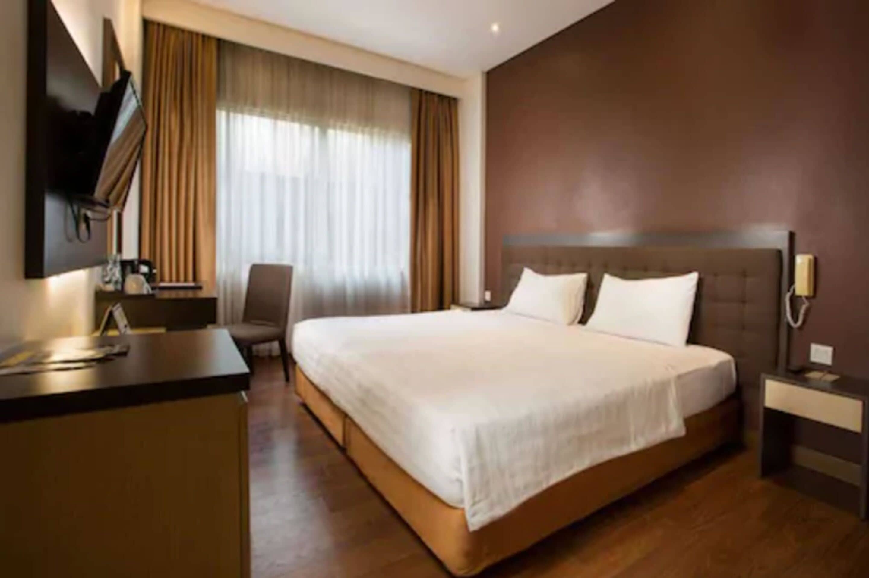 Ein Hotelzimmer mit einem großen Bett, einem Schreibtisch, einem Sessel, einem an der Wand befestigten Fernseher und einem Fenster mit Vorhängen.