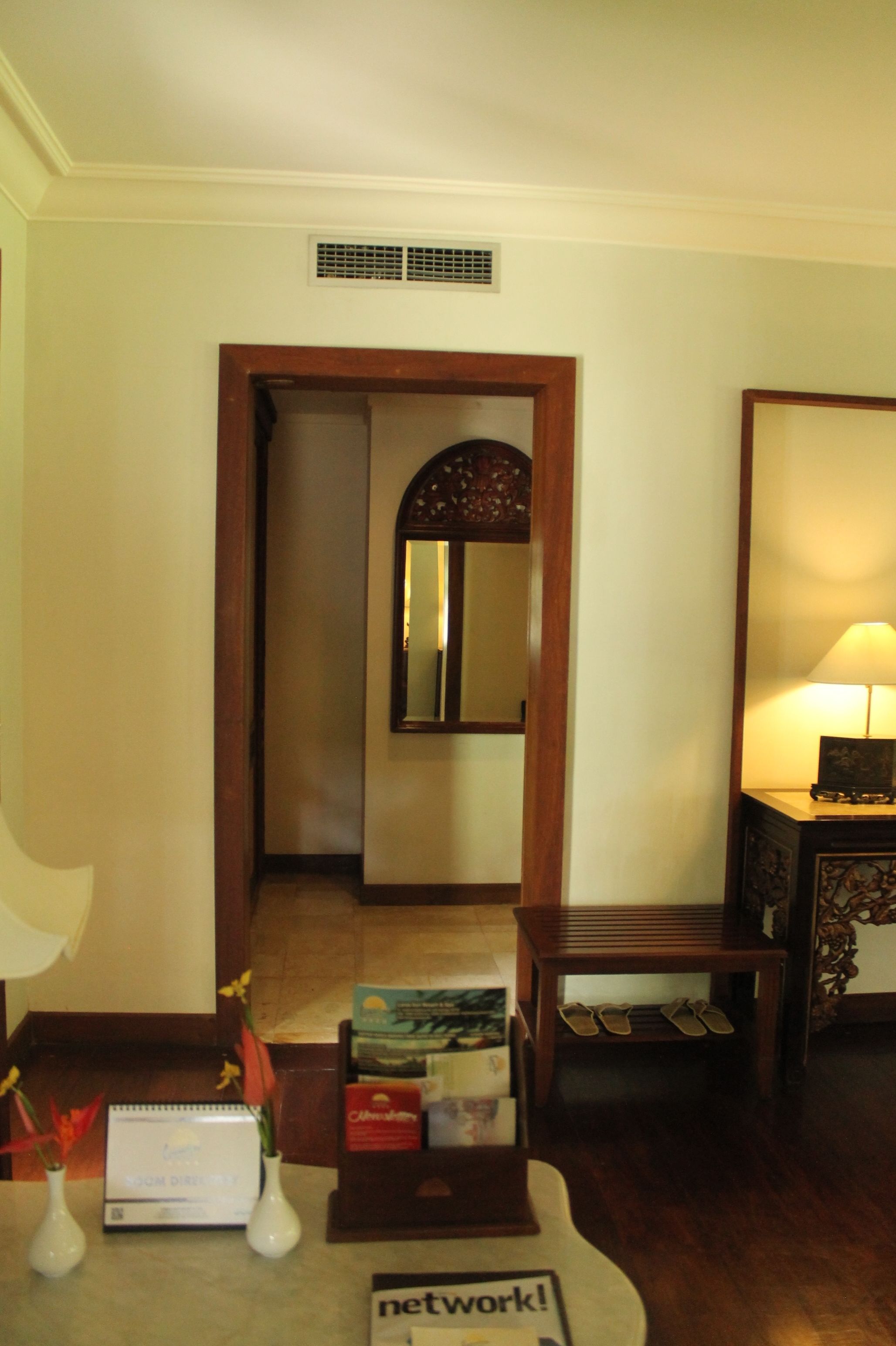 A hotel room with a wooden door, a mirror, a desk with a lamp, and a small table with a book and a vase.