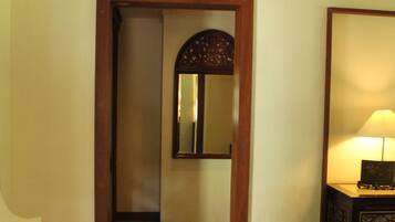 A hotel room with a wooden door, a mirror, a desk with a lamp, and a small table with a book and a vase.