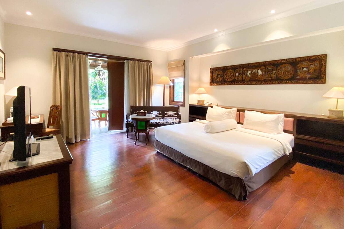 A hotel room with a large bed, a desk with a computer, a chair, and a table. There is a window with curtains and a door leading to an outdoor area.