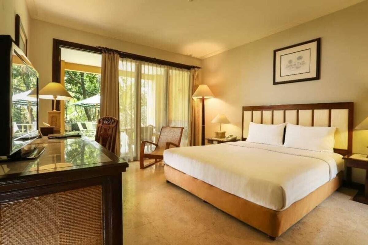 A hotel room with a large bed, a desk with a computer, a chair, and a window with curtains.