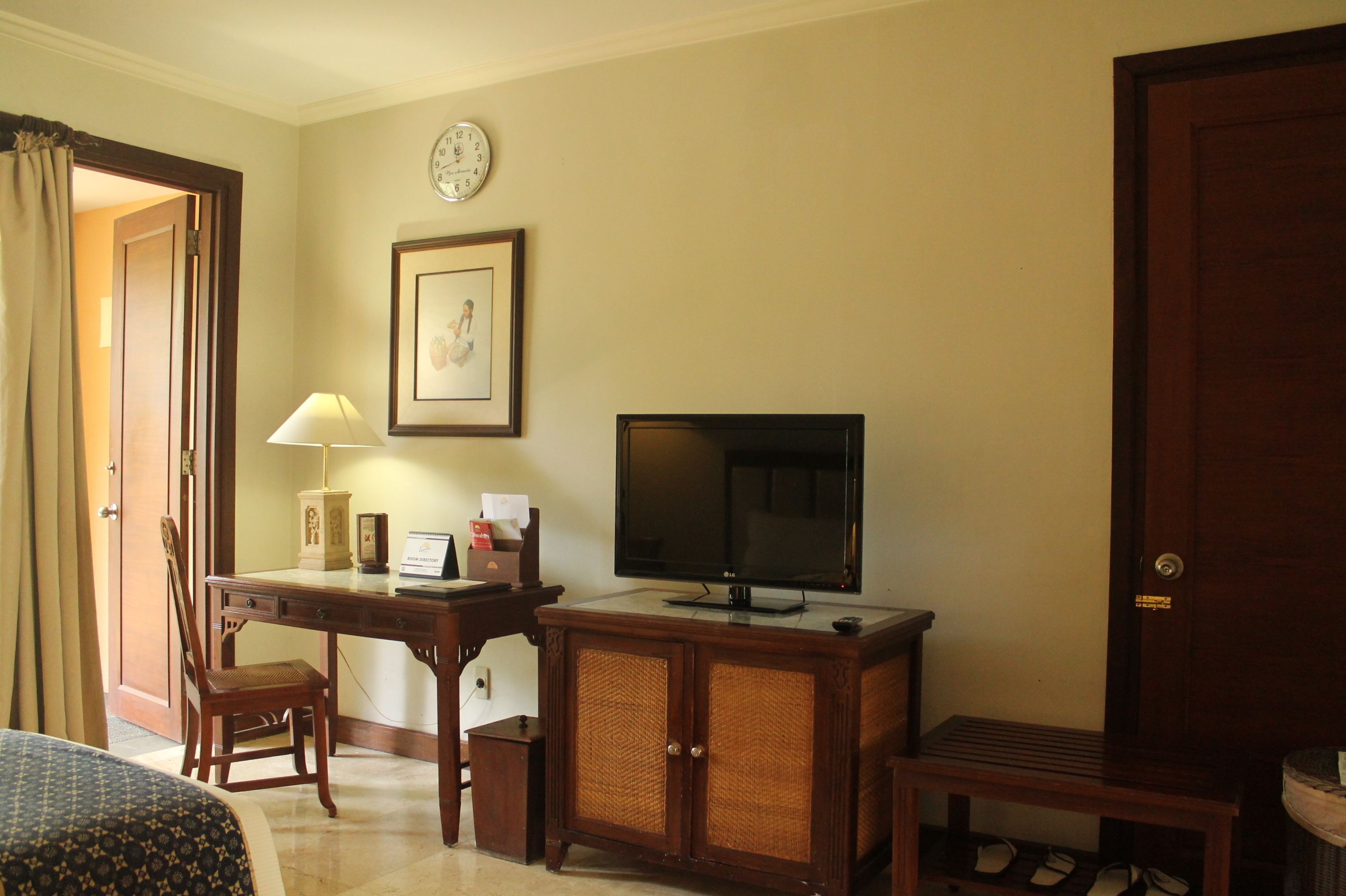 Una habitación de hotel con cama, escritorio con lámpara, televisor, un cuadro en la pared y una puerta.