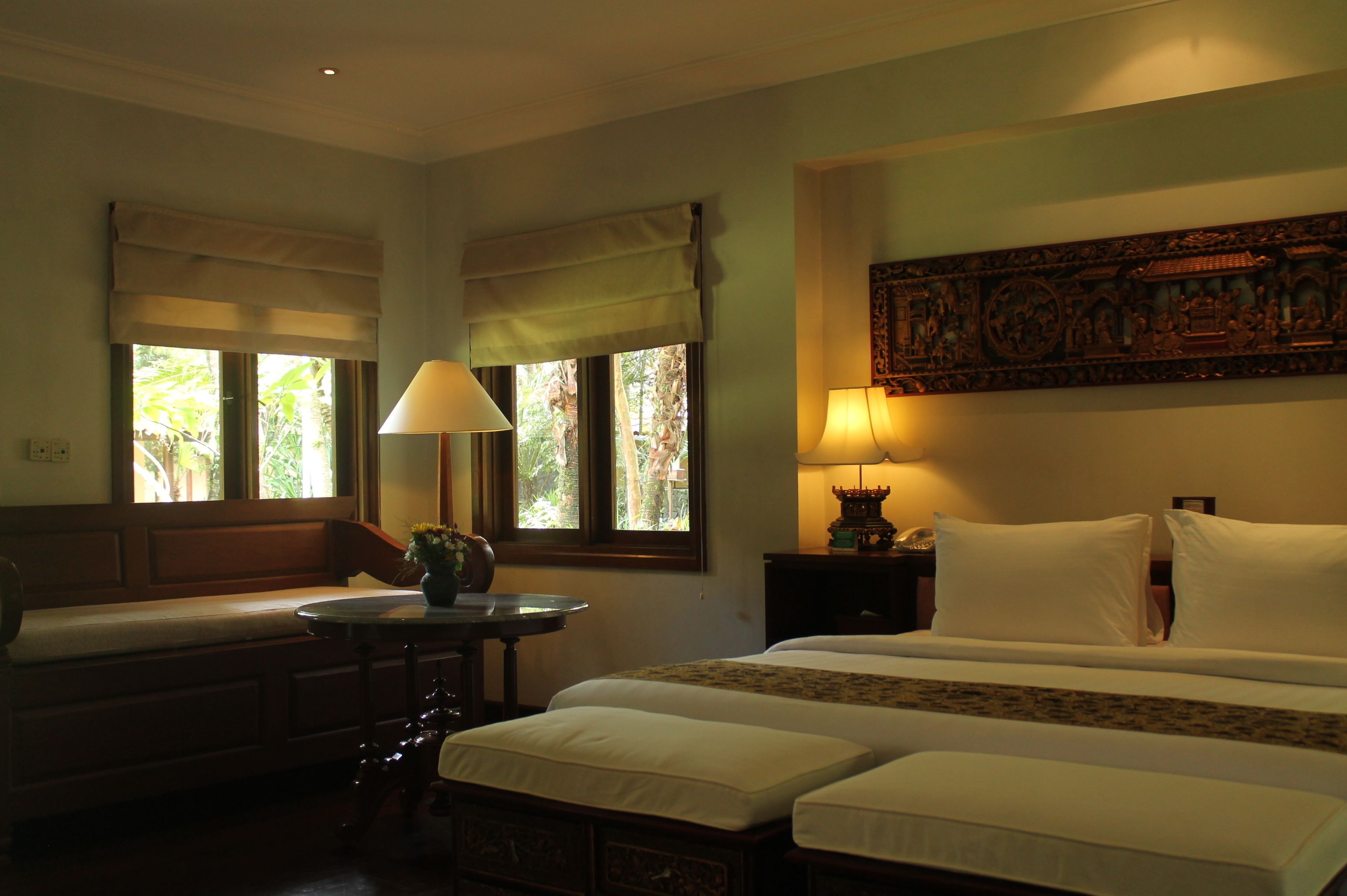 Quarto de hotel com uma cama, duas mesas de cabeceira, um banco e uma janela com vista para o verde.