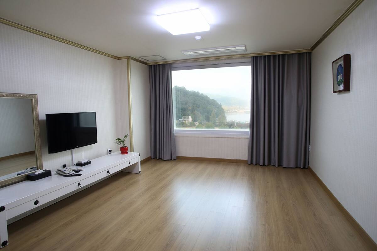 A hotel room with a large window, a flat-screen TV, a bedside table, and a mirror.