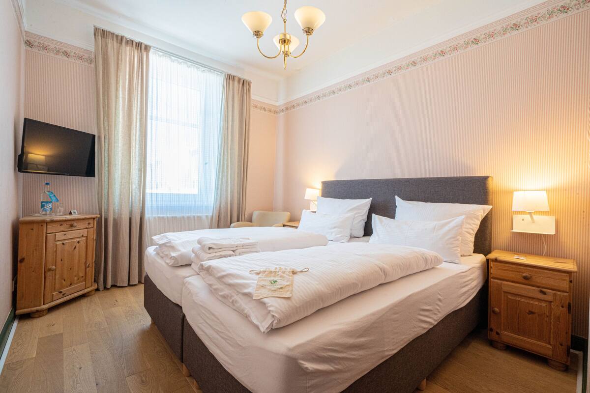 A hotel room with two beds, a television, wooden nightstands, and a chandelier.