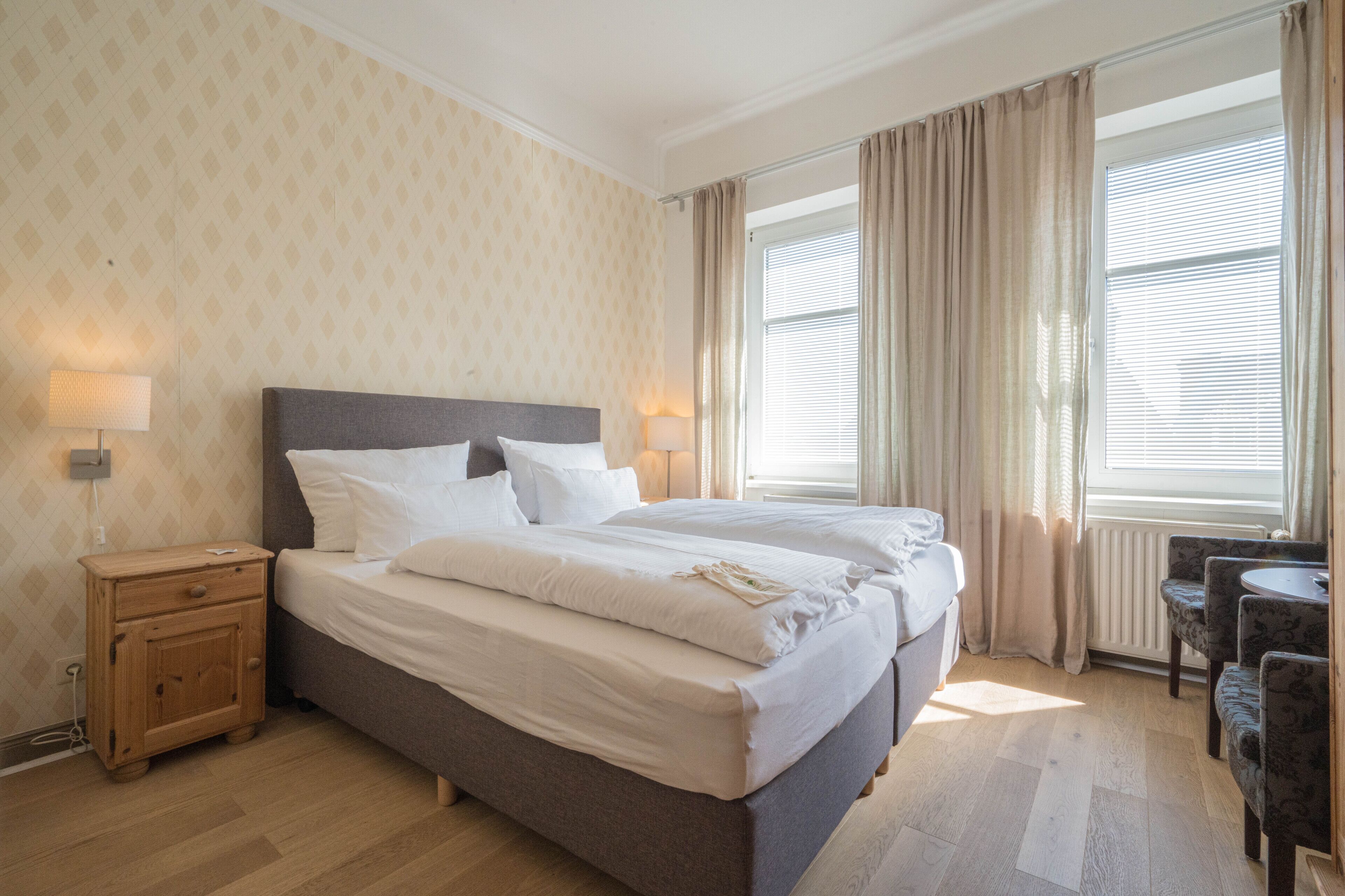 Quarto de hotel com uma cama grande, duas mesas de cabeceira, uma pequena mesa com cadeiras e uma janela com cortinas.