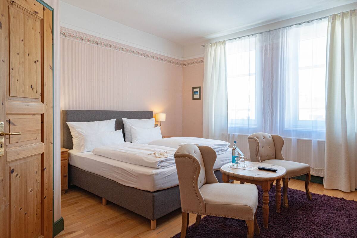 A hotel room with a bed, two chairs, a small table, a nightstand, and a wooden wardrobe.