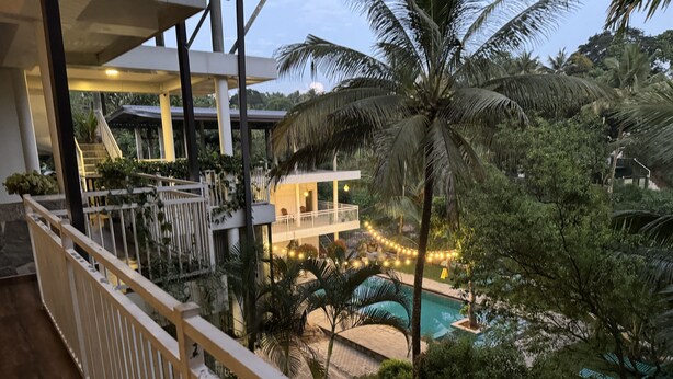 A balcony with a view of a swimming pool, palm trees, and a building with string lights.