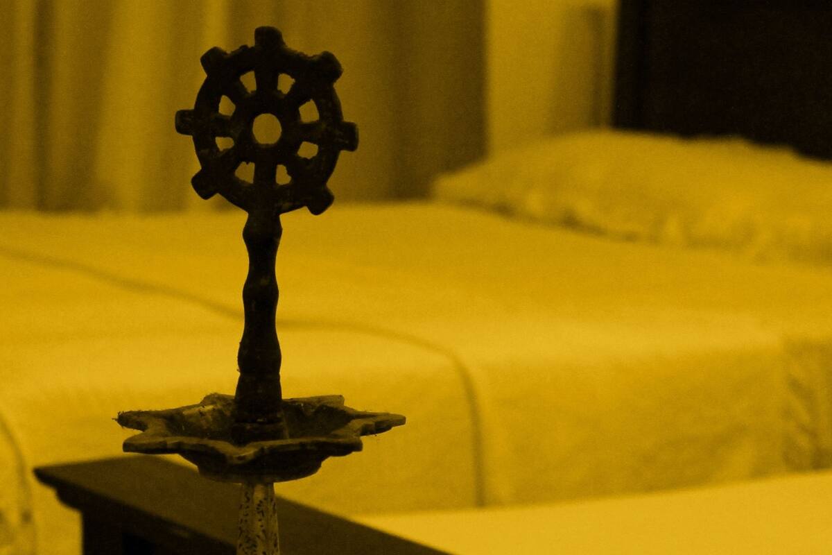 Una habitación tenuemente iluminada con una cama al fondo, una mesa con un objeto decorativo en primer plano y una mesita de noche con una lámpara.