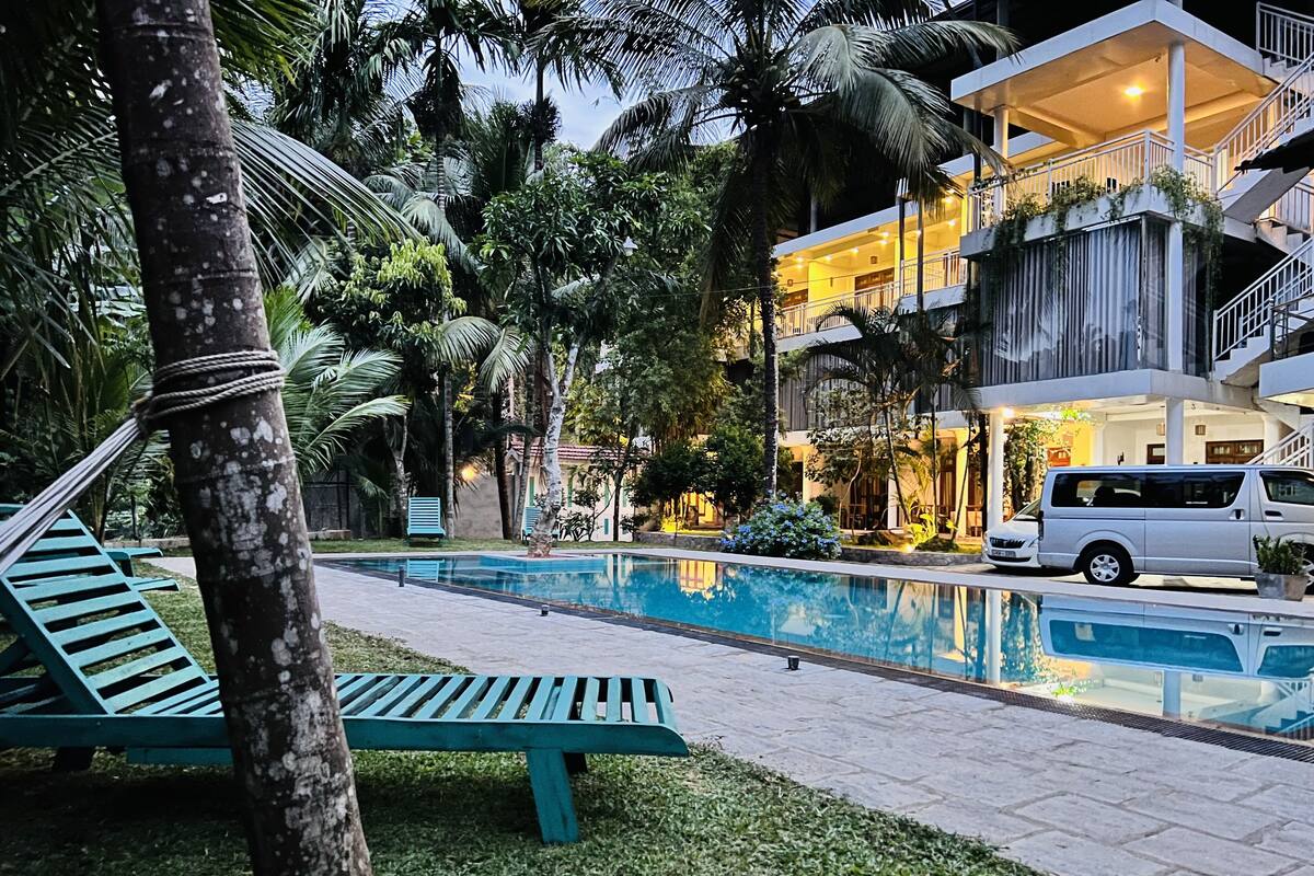 Zona de piscina con sillones reclinables, una camioneta blanca y un edificio con varios balcones e iluminación exterior.
