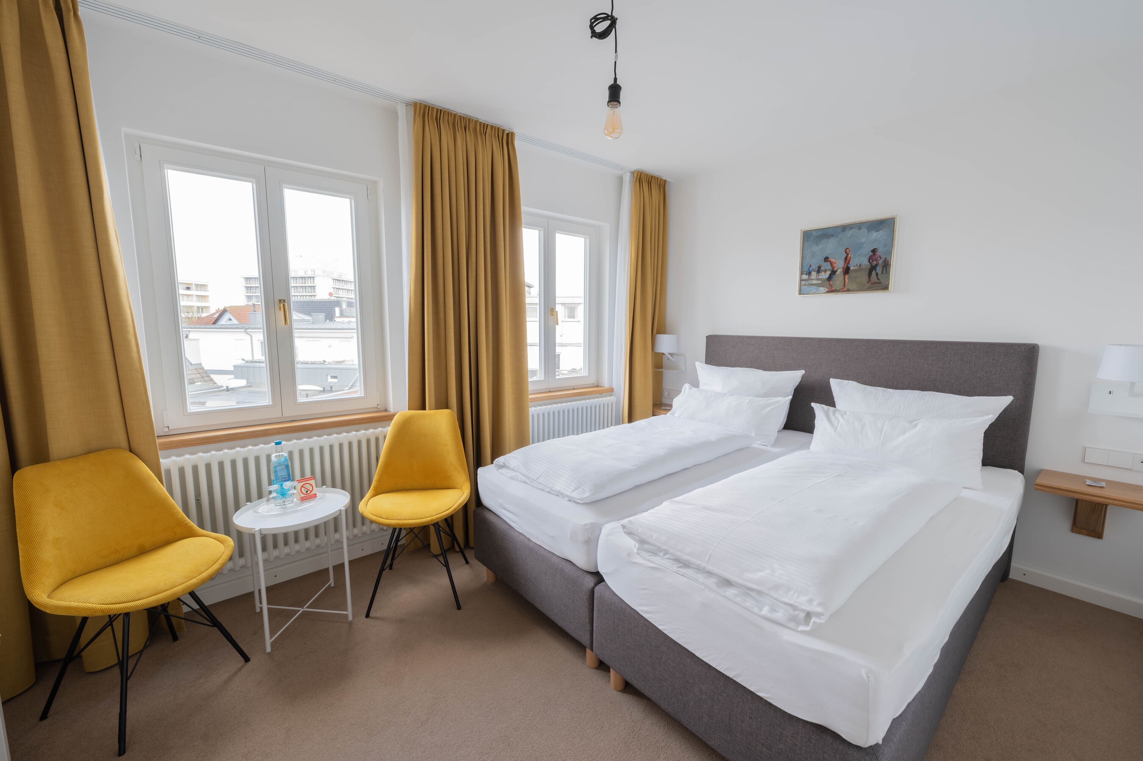 Habitación de hotel con dos camas, una mesita con una silla, un radiador y una ventana con cortinas.