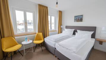 Habitación de hotel con dos camas, una mesita con una silla, un radiador y una ventana con cortinas.