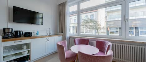 A modern kitchen with white cabinets, a flat-screen TV, a dining table with pink chairs, and a large window with a view of buildings.