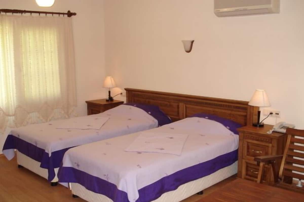 A hotel room with two single beds, wooden headboards, bedside tables with lamps, a small wooden table, and a window with curtains.