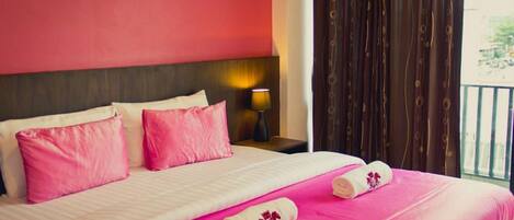 A hotel room with a bed, pink bedding, two pillows, a nightstand with a lamp, and a window with curtains.