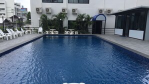 A swimming pool with white lounge chairs and tables, surrounded by air conditioning units and a building with a blue awning.