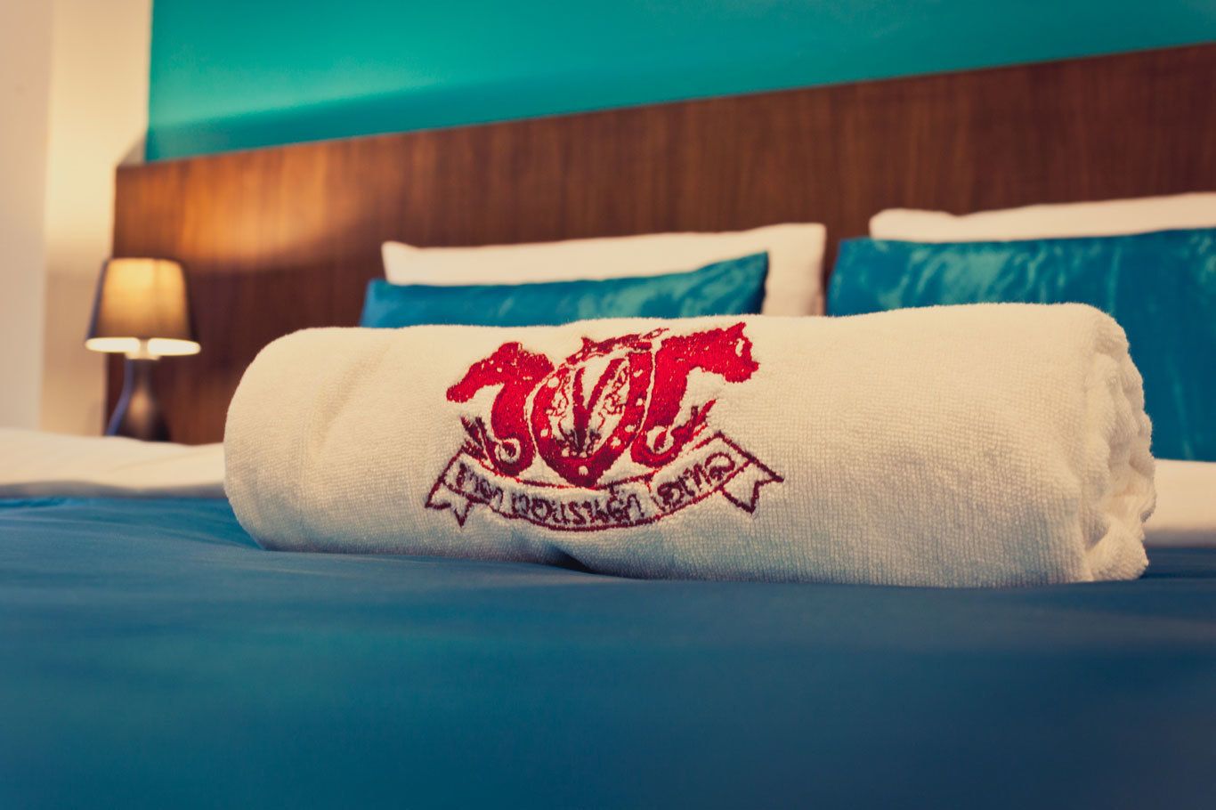 A neatly made bed with a rolled white towel featuring a red logo and text, blue bedding, and a wooden headboard.