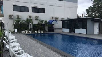 Una piscina con sillas y mesas de descanso blancas, unidades de aire acondicionado y un edificio con ventanas grandes y un toldo azul.