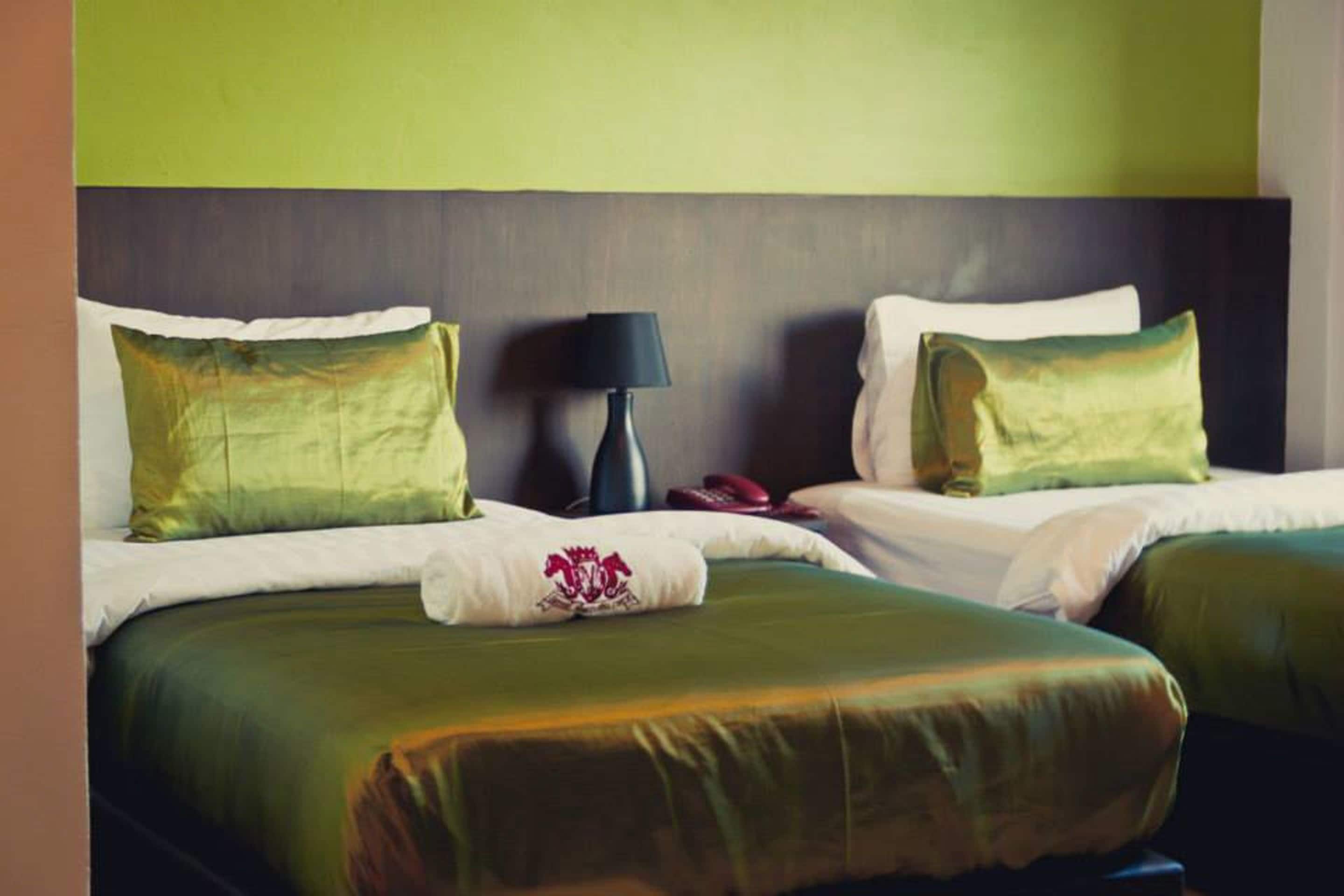 Una habitación de hotel con dos camas, cada una con ropa de cama verde y almohadas blancas, un cabecero oscuro y una lámpara de noche.
