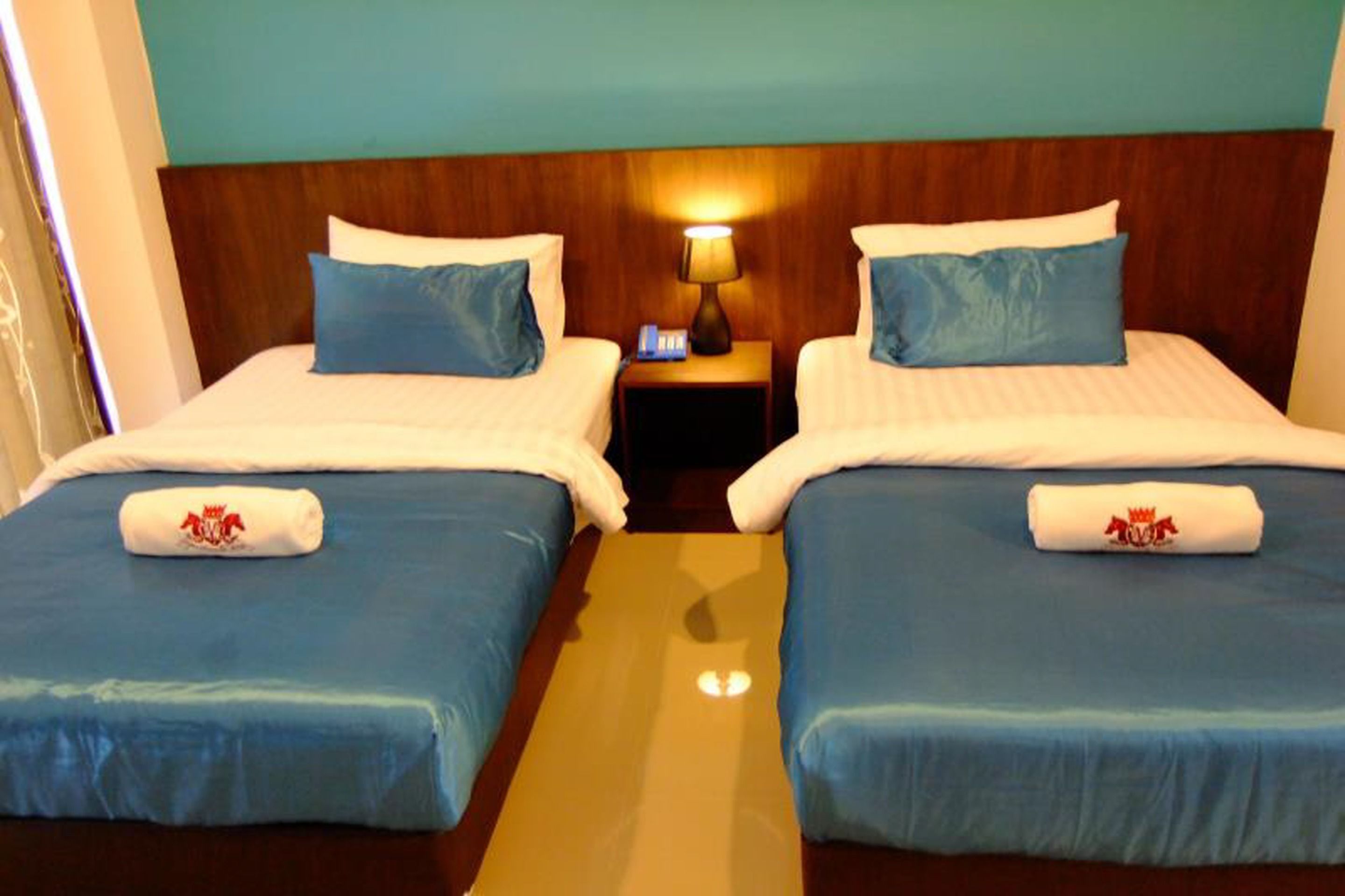 Habitación de hotel con dos camas, cada una con cubrecama y almohadas azules, y una mesita de noche con lámpara entre ellas.