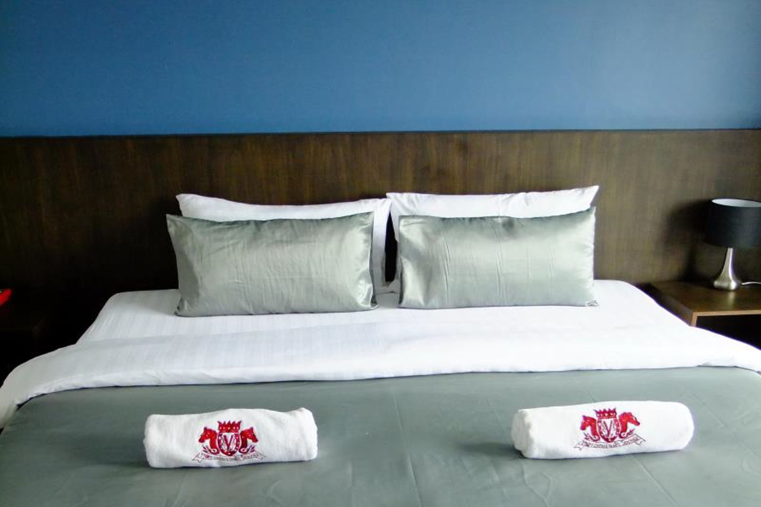 A neatly made bed with two pillows and two towels with a crest logo, a wooden headboard, and a nightstand with a lamp.