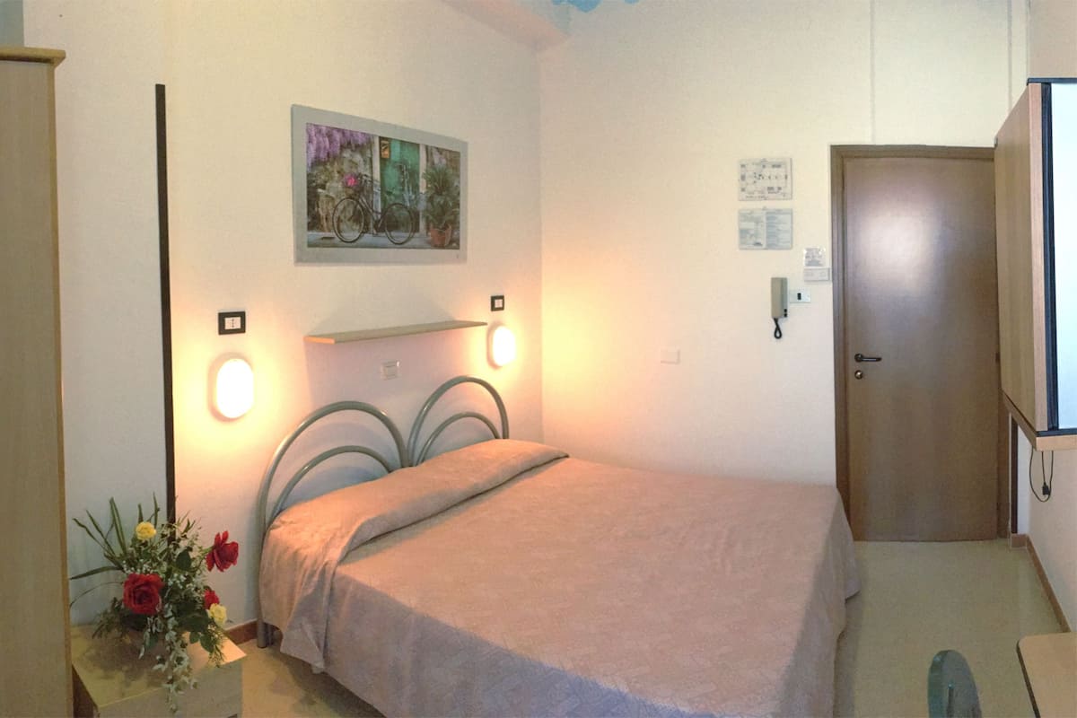 A bedroom with a bed, a nightstand with a vase of flowers, a door, and a wall-mounted shelf with a picture.