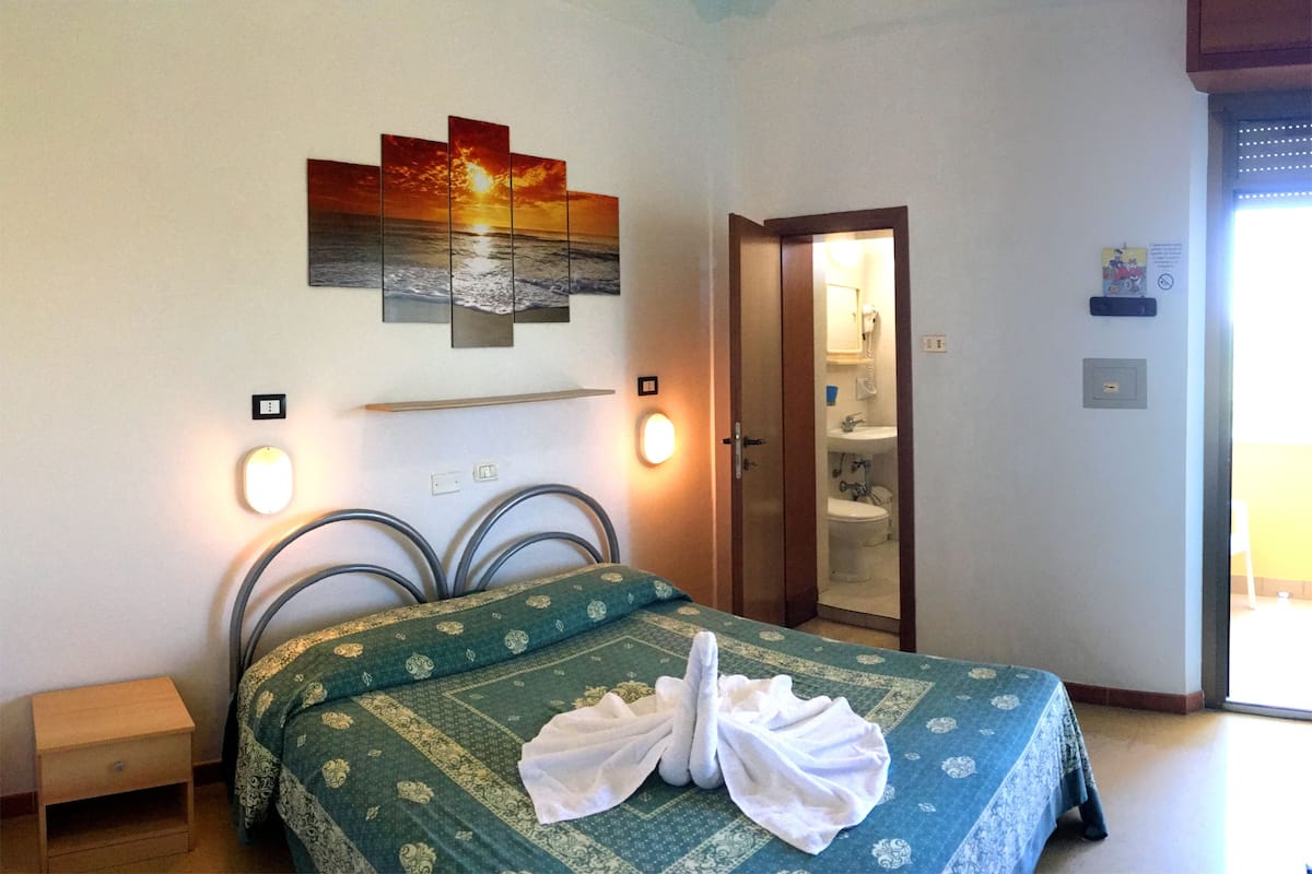 A hotel room with a bed, a nightstand, a bathroom with a shower, and a door leading to another room.