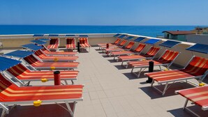 Un salón en la azotea con hileras de sillones a rayas y sombrillas, con vistas al mar azul y despejado.