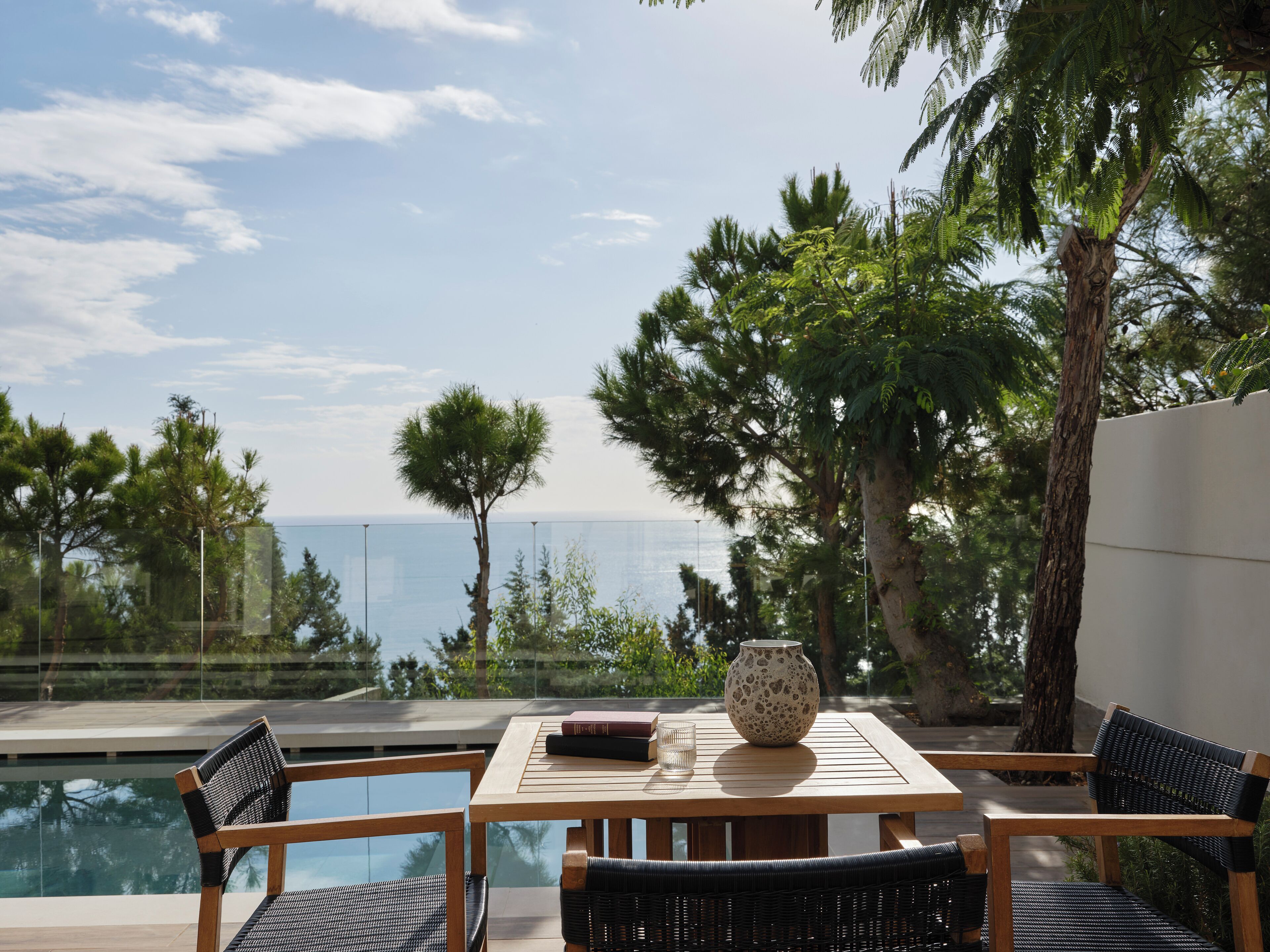 A wooden table with chairs, a vase, and books, overlooking a swimming pool and a view of the sea.