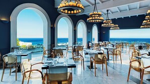 Un restaurant doté de nombreuses tables et chaises, de grandes fenêtres offrant une vue sur l’océan et de lustres suspendus.