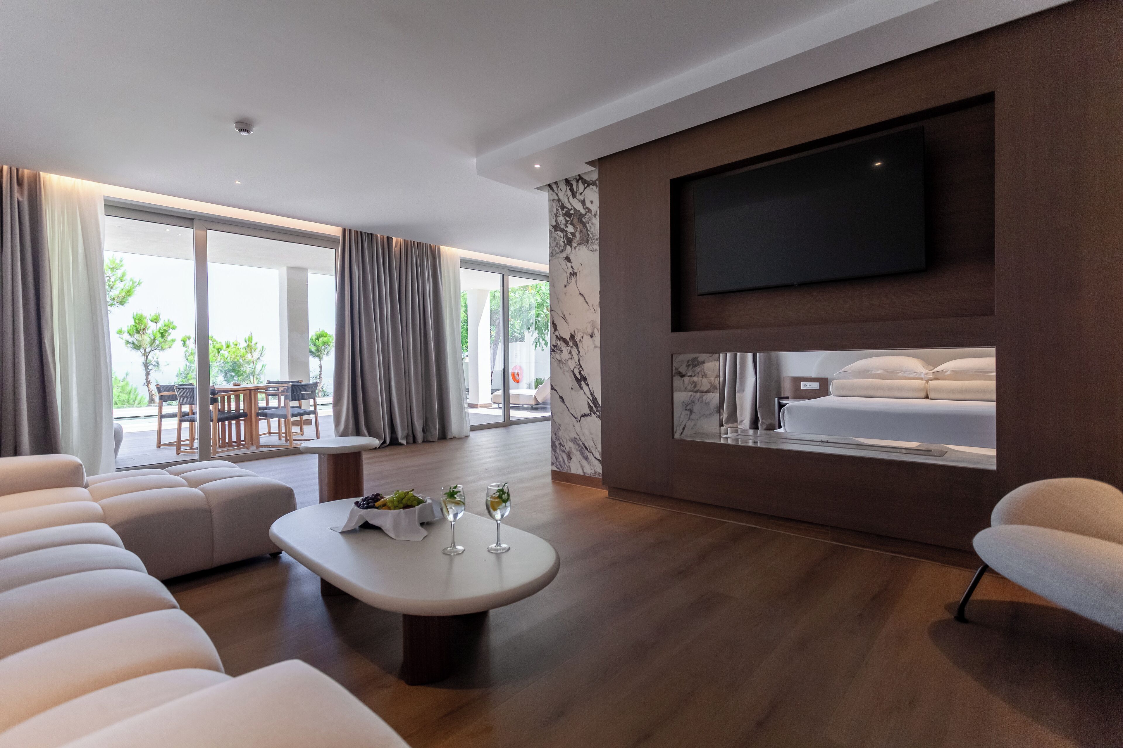 Ein modernes Hotelzimmer mit großem Flachbildfernseher an der Wand, einer gemütlichen Couch, einer Essecke mit Tisch und Stühlen und einem großen Fenster mit Vorhängen.