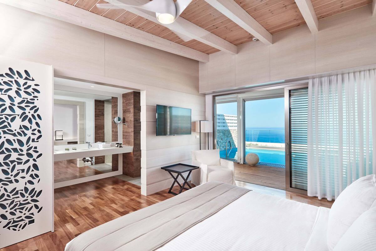 Une chambre moderne avec un plafond en bois, un grand lit, une salle de bain avec un miroir et un lavabo, et un balcon avec vue sur la piscine.