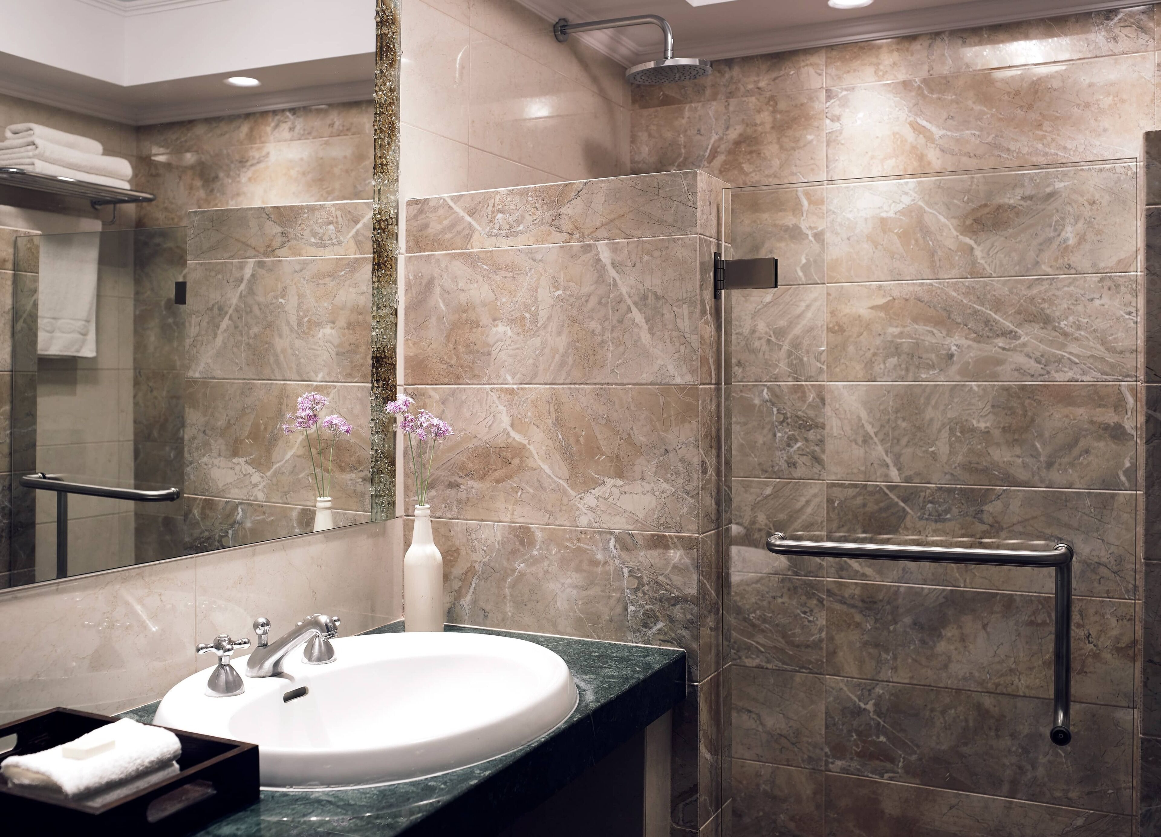 A modern bathroom with a marble sink, a glass shower enclosure, and a handrail.