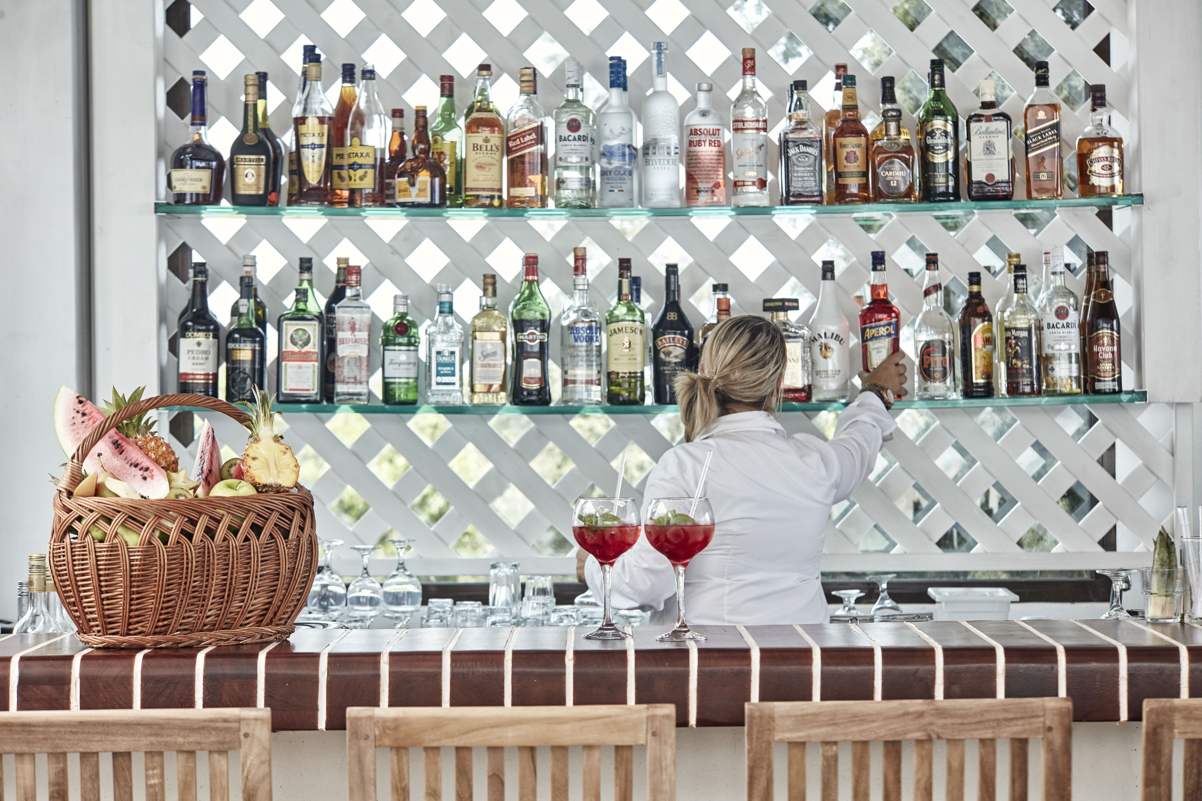 Un coin bar avec une variété de bouteilles d’alcool exposées sur des étagères, un panier en osier rempli de fruits et deux verres contenant une boisson rouge.