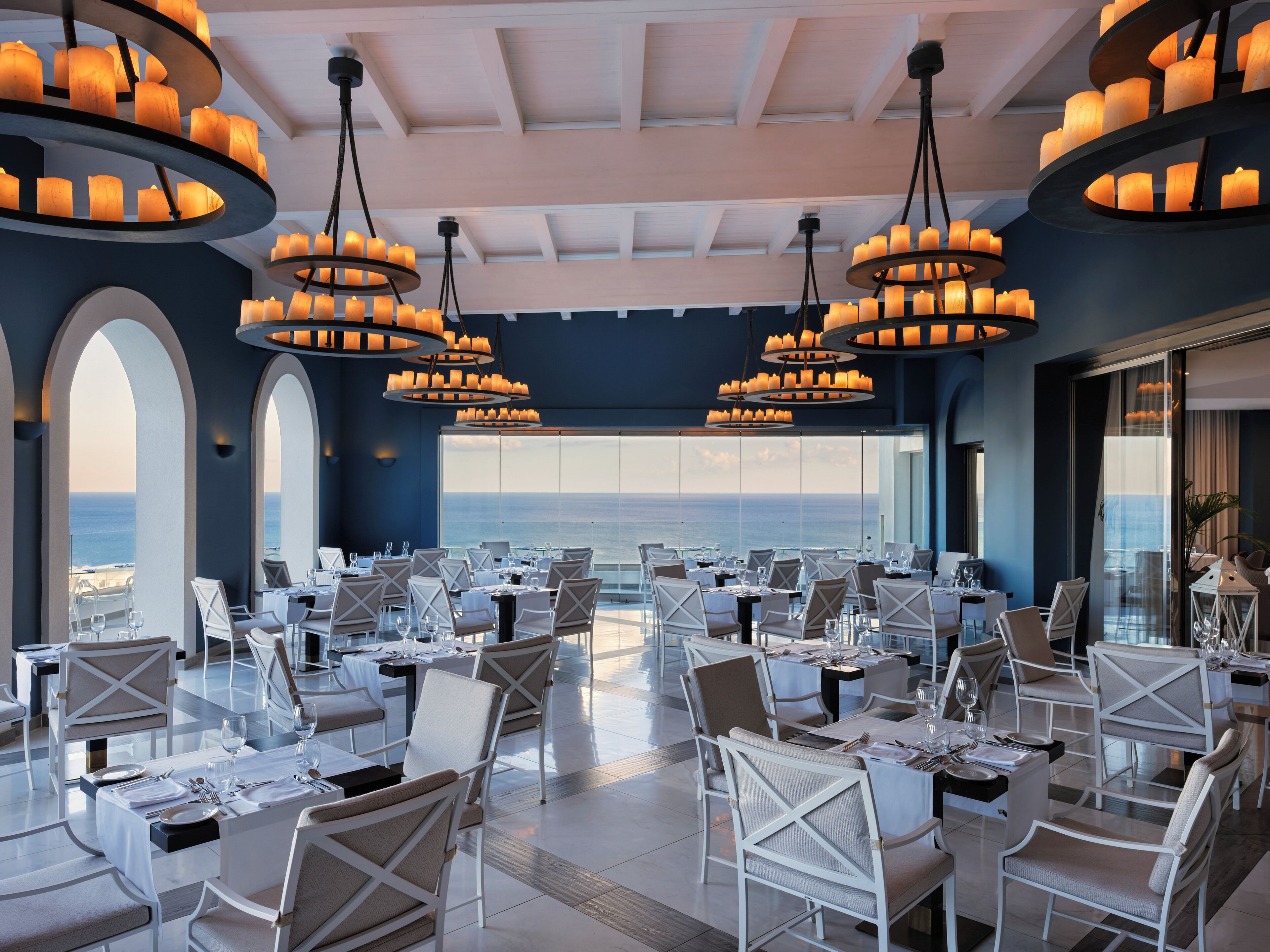 Une salle à manger spacieuse, dotée de nombreuses tables et chaises, de grandes fenêtres offrant une vue sur l’océan et de magnifiques lustres.