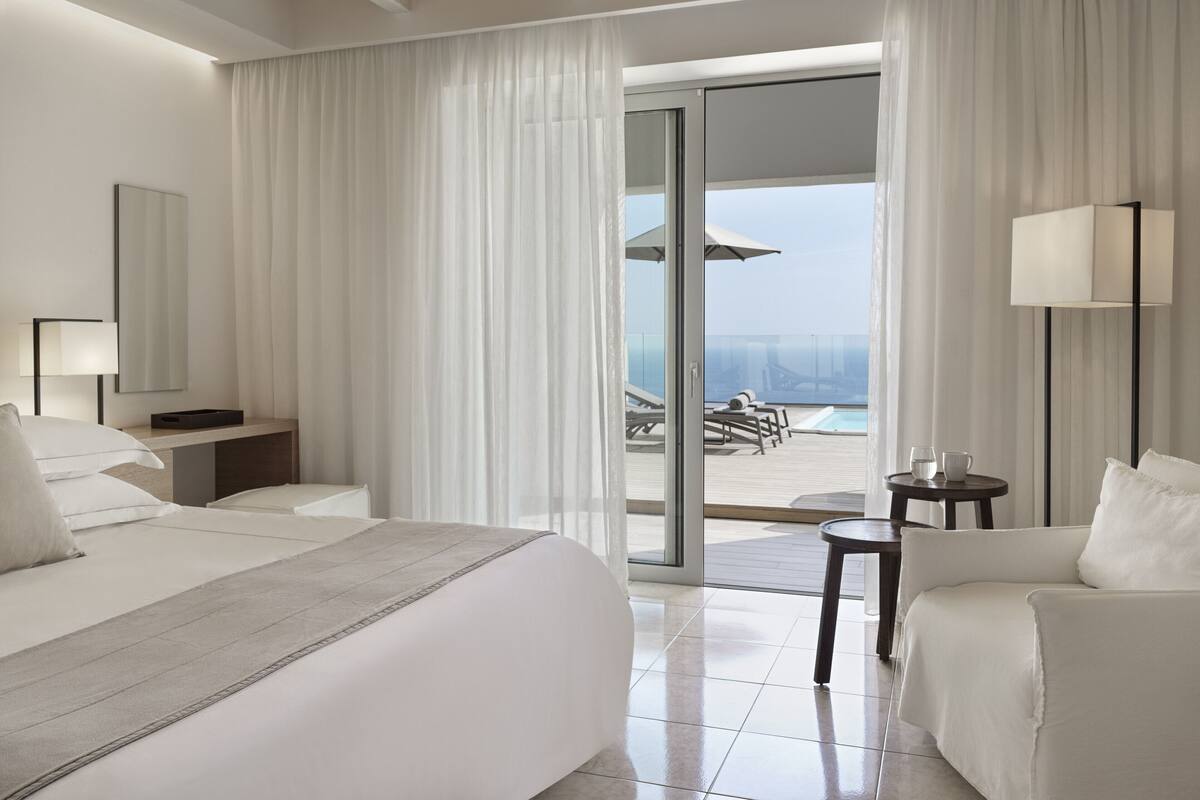 Une chambre d’hôtel moderne dotée d’un grand lit, d’un coin salon avec un fauteuil et une petite table, d’un bureau avec une lampe, et offrant une vue sur une piscine et une plage, le tout visible à travers une porte vitrée.