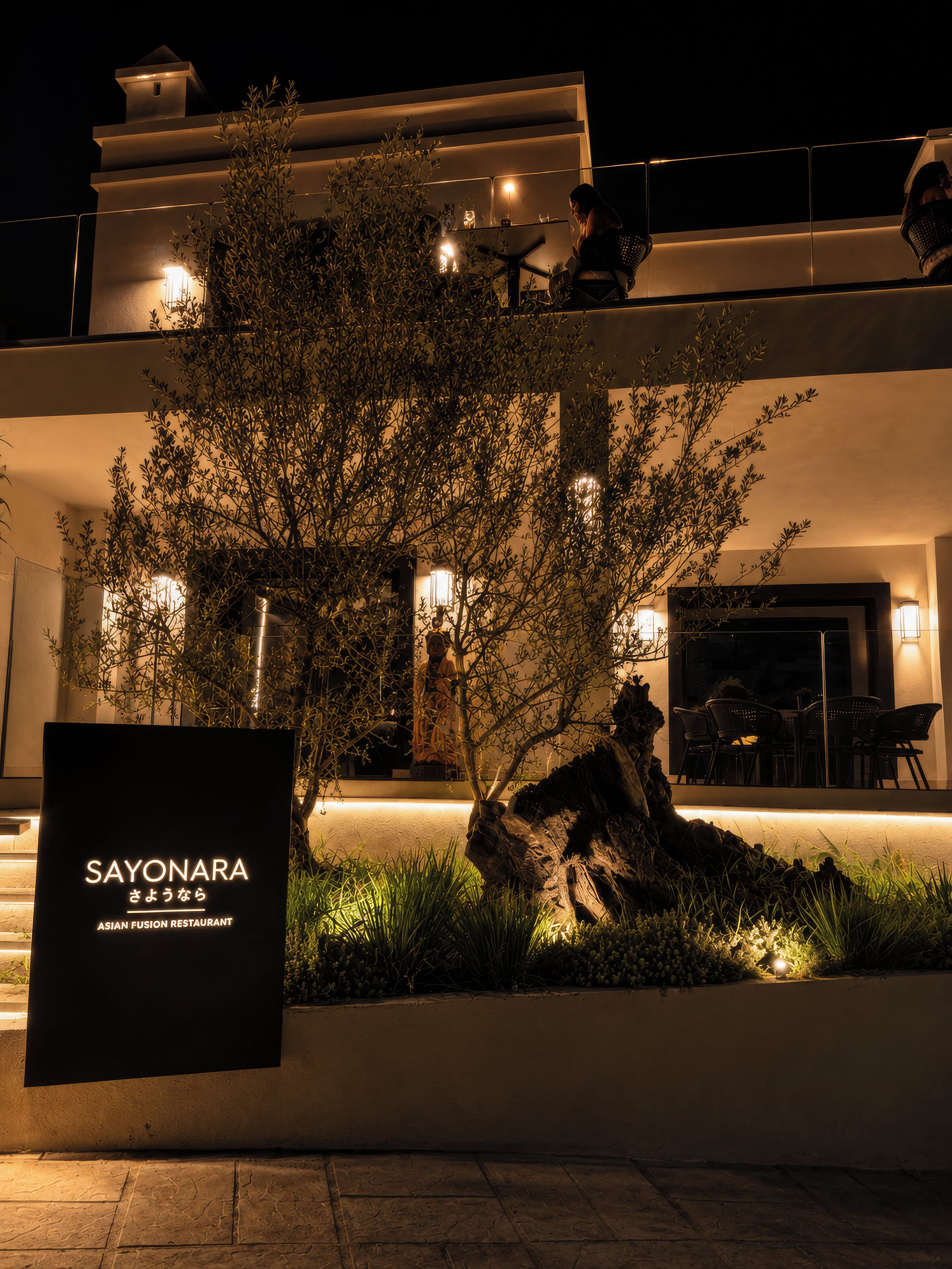 Un edificio moderno con un letrero que dice 'SAYONARA Asian Fusion Restaurant' y mesas al aire libre.
