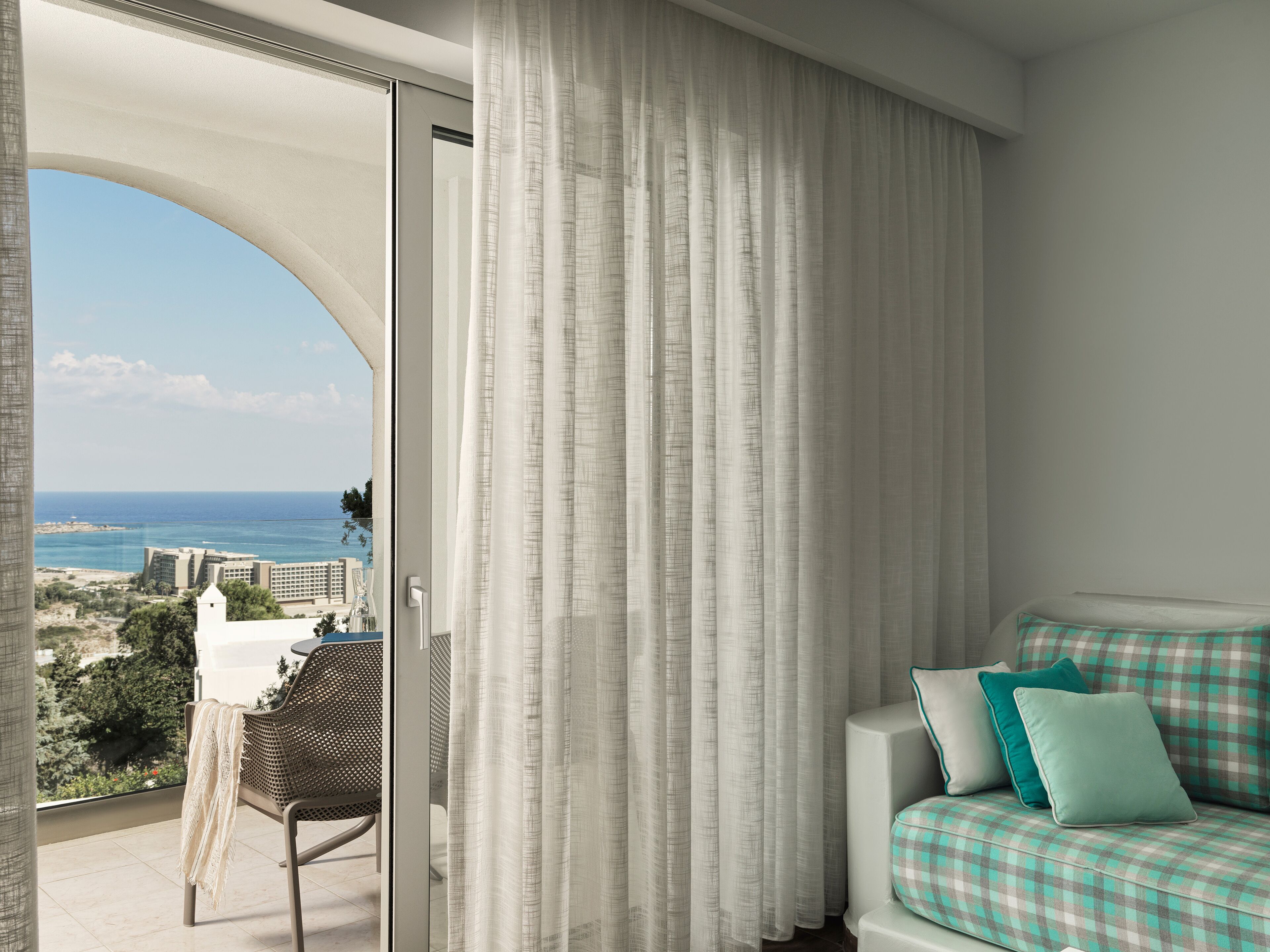 A balcony with a view of the sea and buildings, a wicker chair, a sofa with teal cushions, and a white curtain.