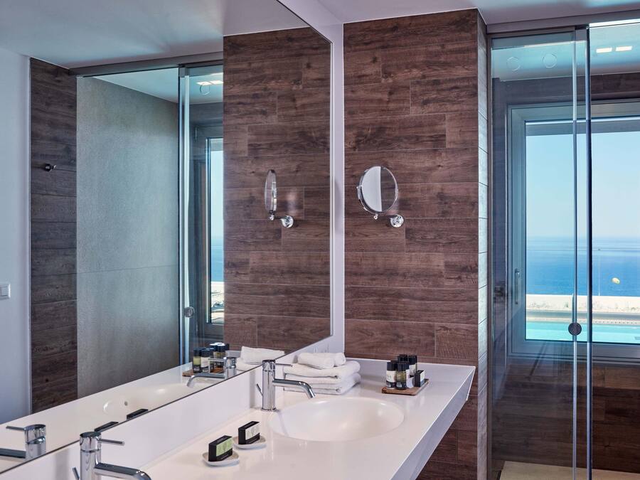 Un baño moderno con un espejo grande, dos lavamanos y vistas a la playa a través de una puerta de cristal.