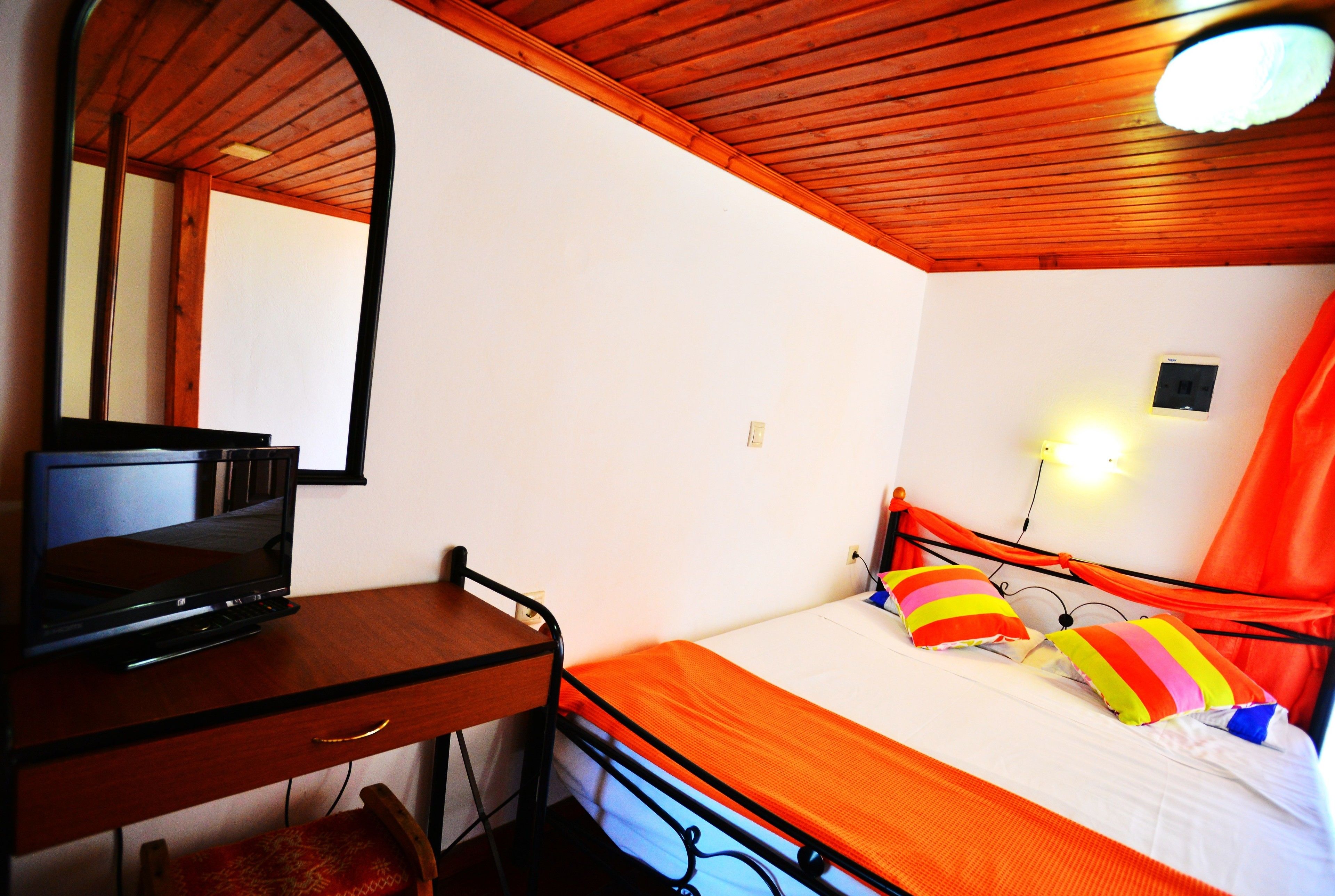 A bedroom with a wooden ceiling, a bed with colorful pillows, a bedside table with a television, and a mirror.