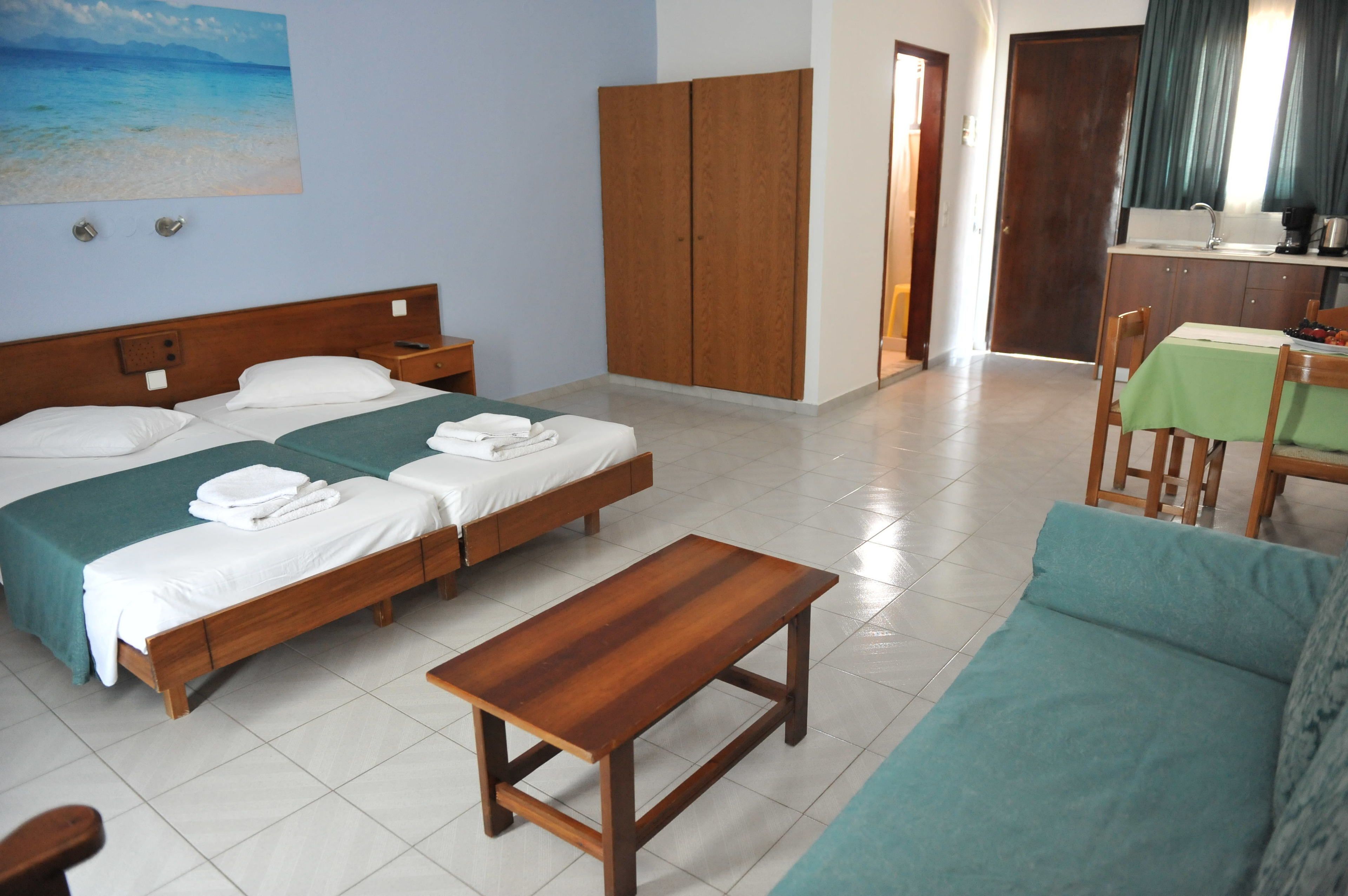 Habitación de hotel con dos camas, una mesa de madera, un sofá verde y una zona de cocina al fondo.