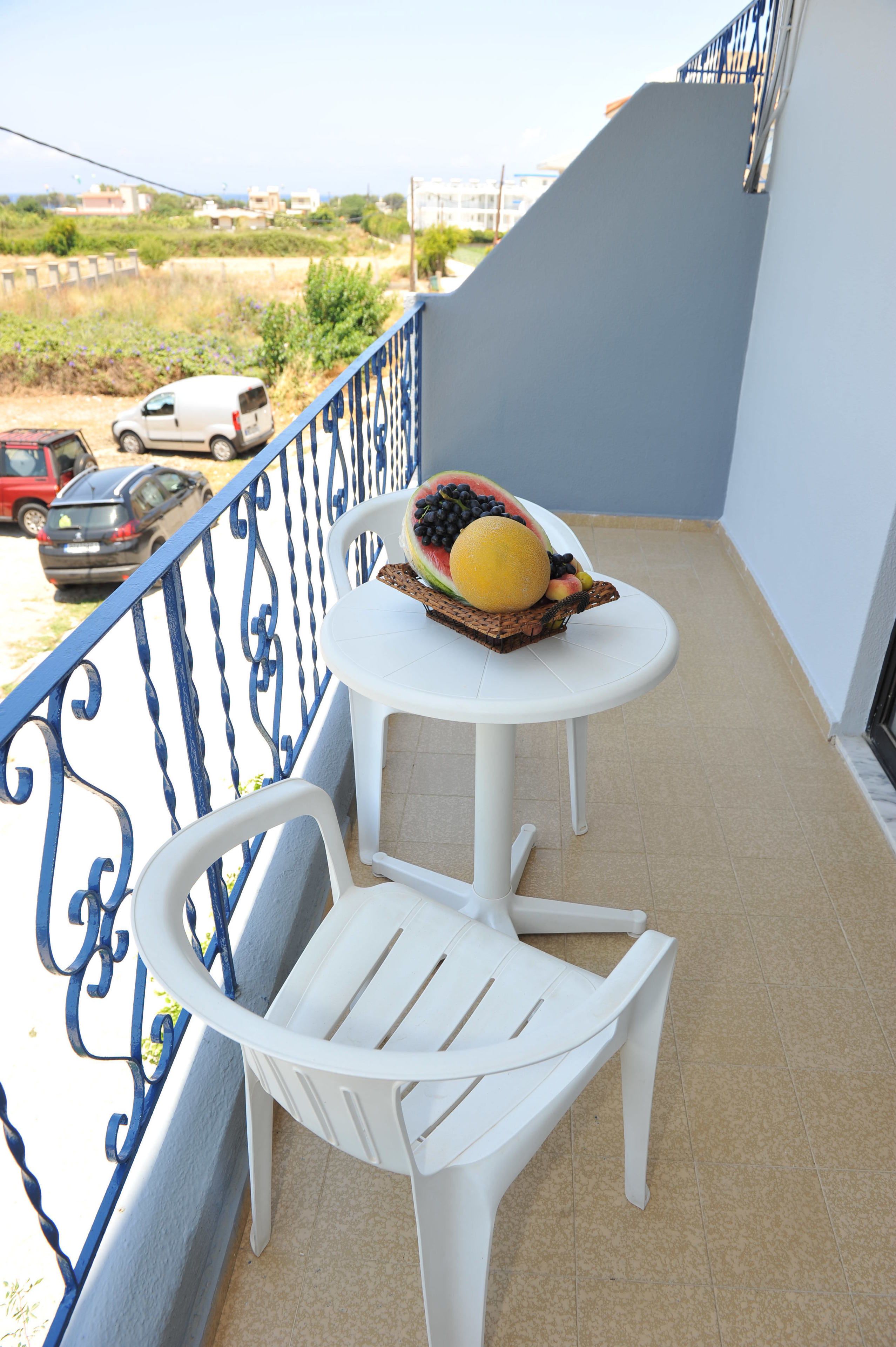 Un balcon avec une chaise et une table blanches, un panier de fruits contenant une pastèque, des myrtilles et une orange, et une vue sur les voitures garées et les bâtiments en arrière-plan.