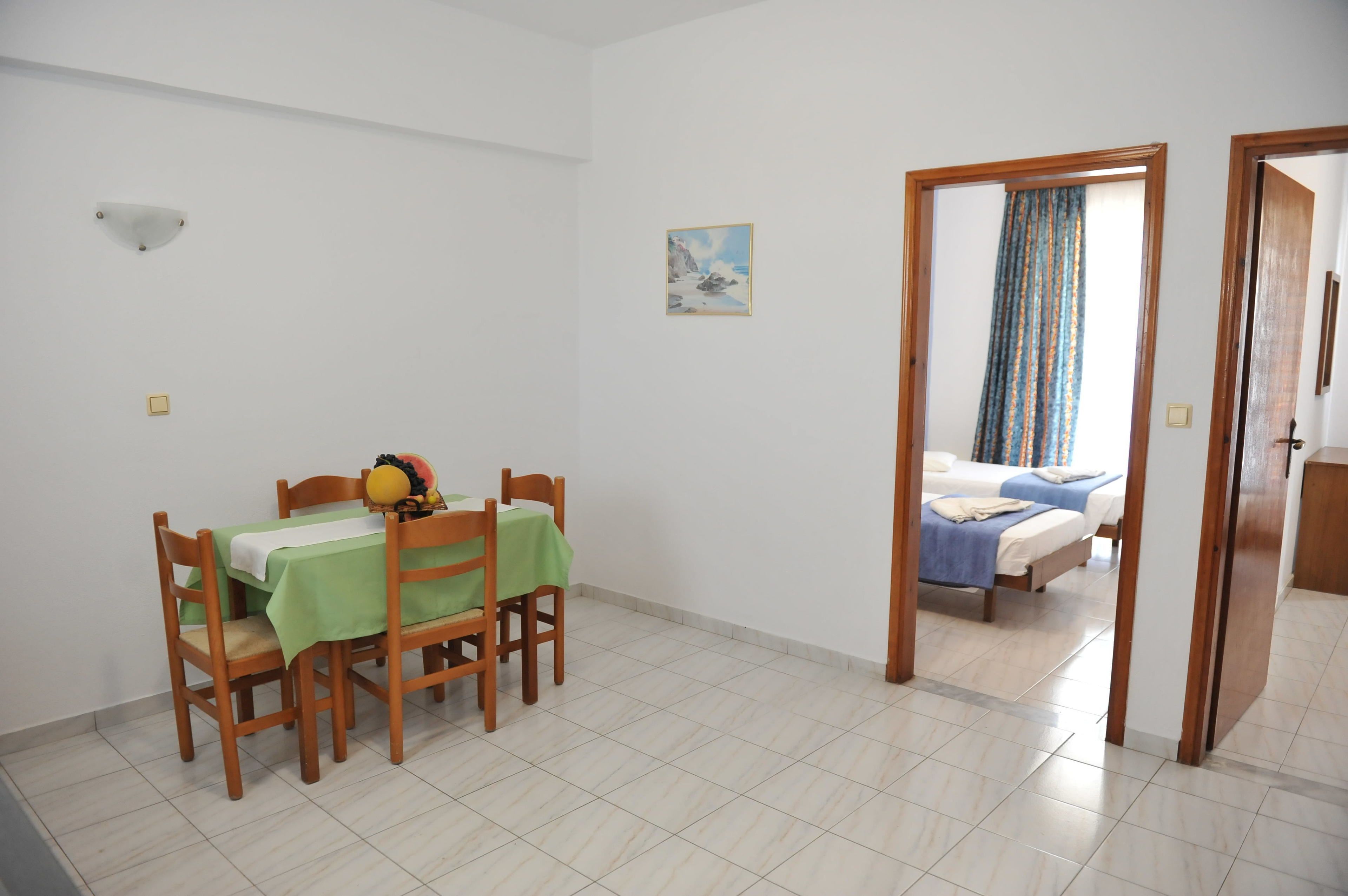 A room with a dining table set with chairs, a bed with a blue blanket, a painting on the wall, and a door leading to another room.