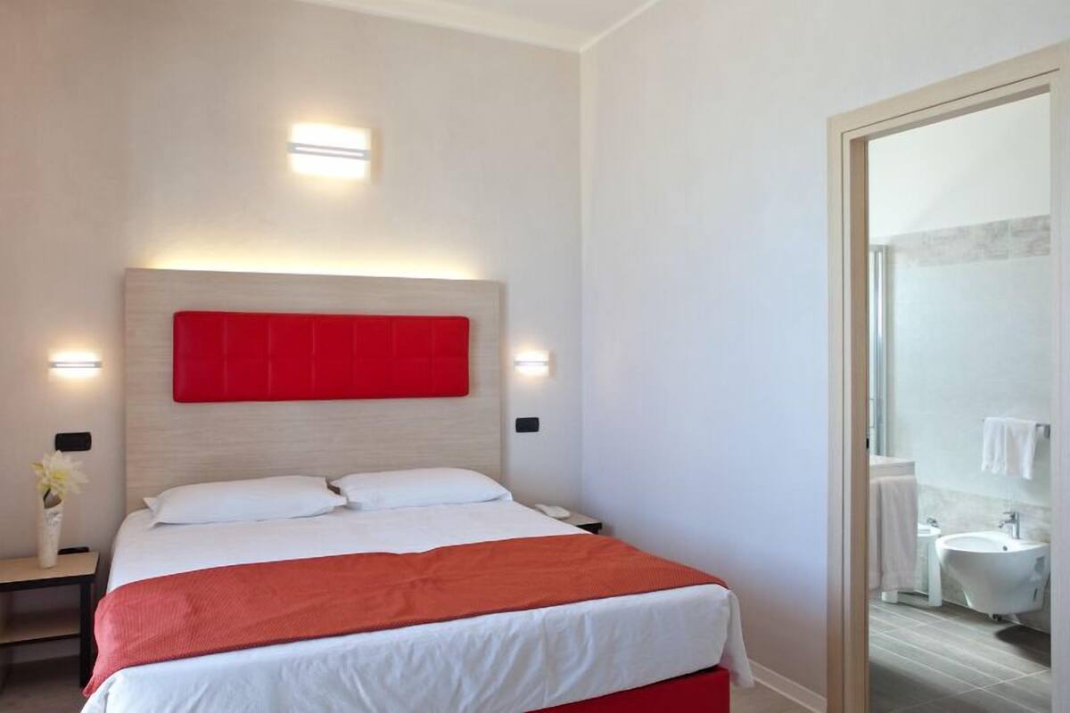 Ein modernes Hotelzimmer mit einem großen Bett, einer roten Akzentwand und einem durch eine geöffnete Tür einsehbaren Badezimmer.