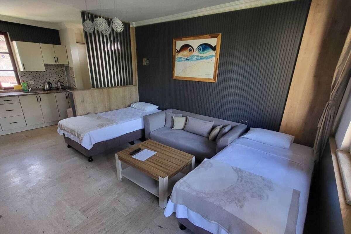 A modern hotel room with two single beds, a sofa bed, a small wooden table, and a kitchen area with cabinets and a countertop.